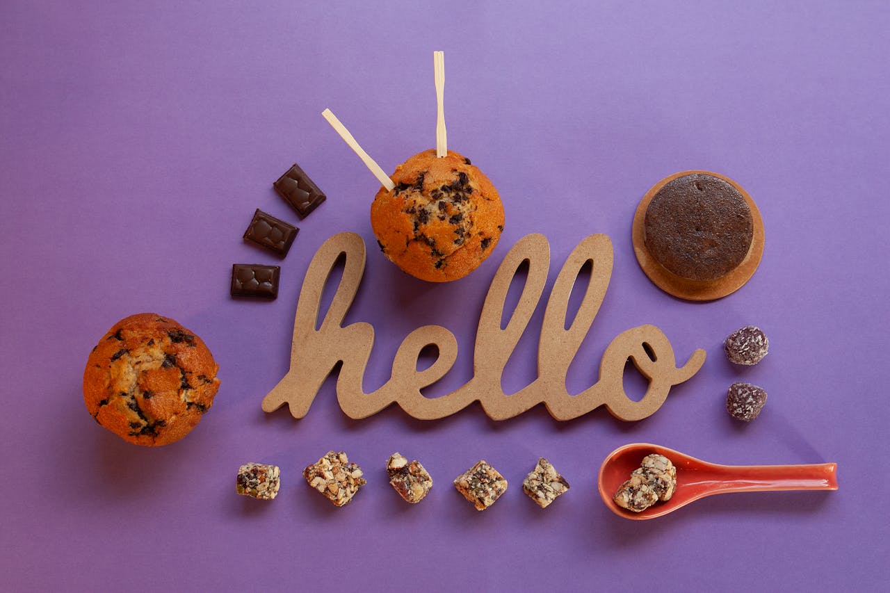 A top view of assorted chocolates and muffins with a 'Hello' sign on a purple surface.